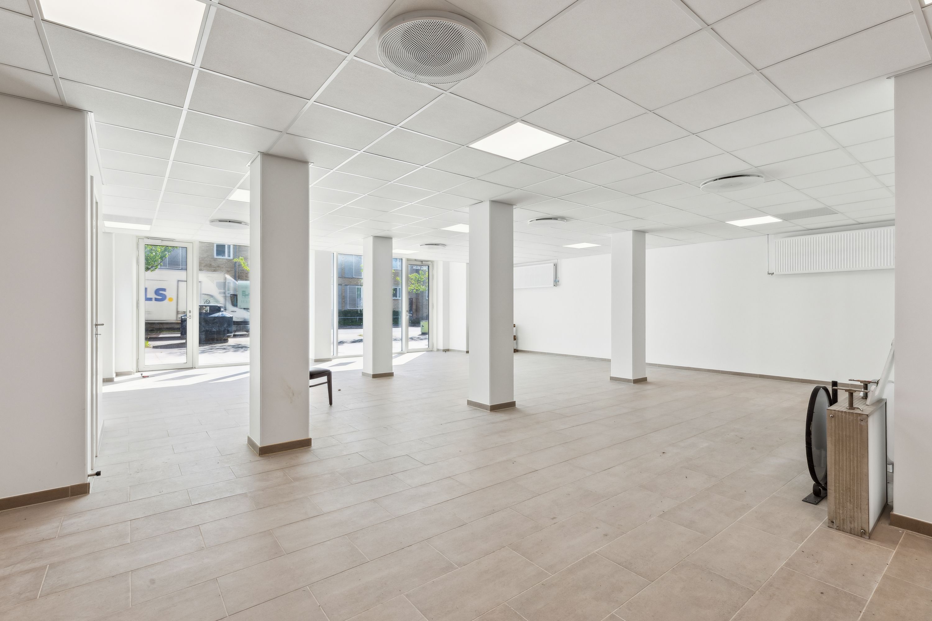 En tom og ren møbleret kontorfællesskab med hvidt loft, hvide vægge, lysegul gulvbelægning, glasdøre og store vinduer