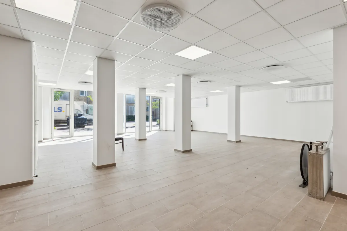 En tom og ren møbleret kontorfællesskab med hvidt loft, hvide vægge, lysegul gulvbelægning, glasdøre og store vinduer