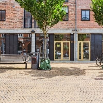 Parkplads med bænk og skraldespand bag en højt muret bygning, træer og en cykelparkeringsplads