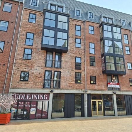 Bygning med røde mursten og glasvinduer, guldfarvet indgangsdør og rødt banner med teksten Udleijing, rødt potteplante og to