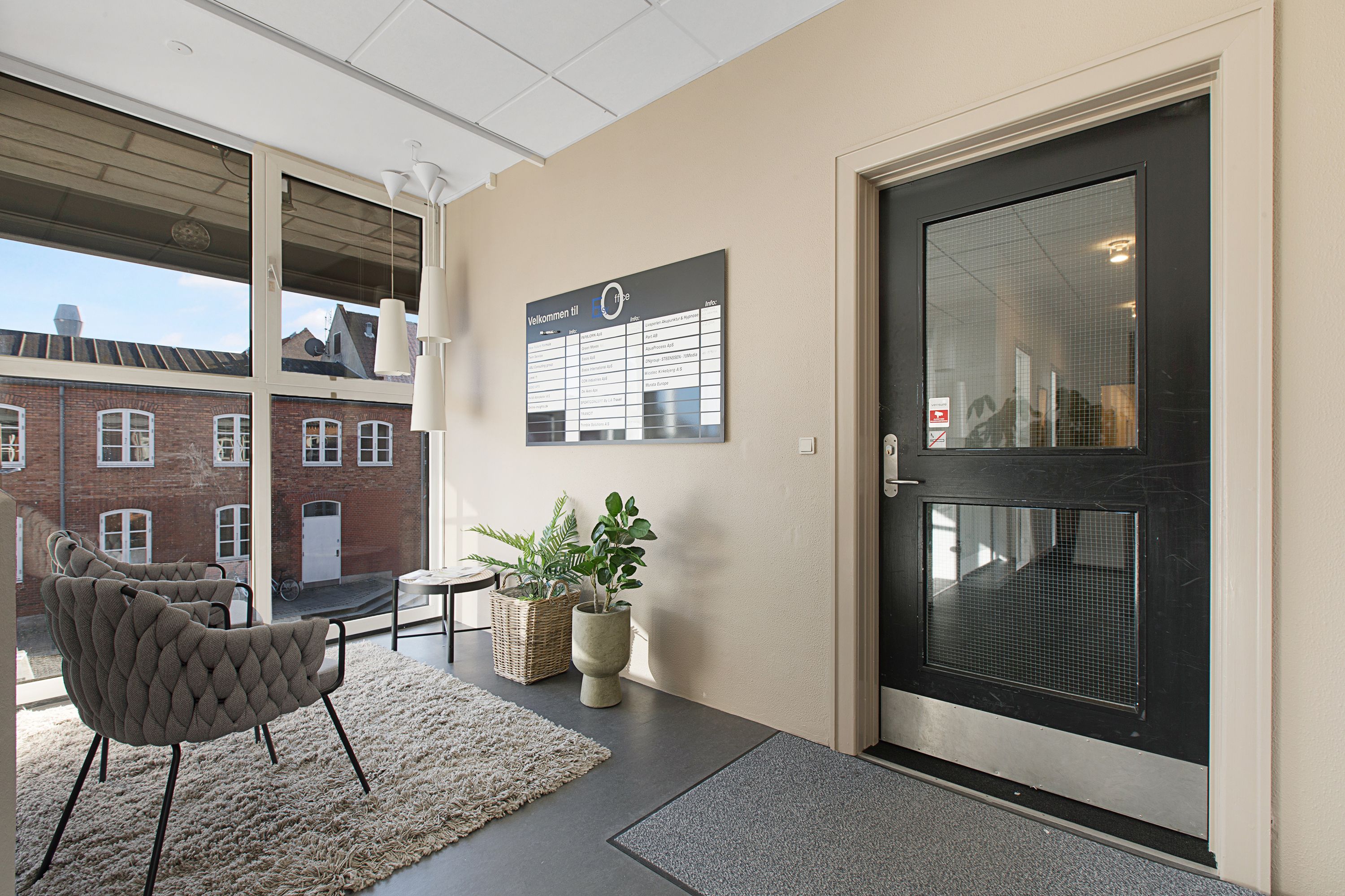 Kontor med indgangsdør og plakat. Et område med to stole, plante og tæppe. Vindue med udsigt til bygning. Lamper hængende