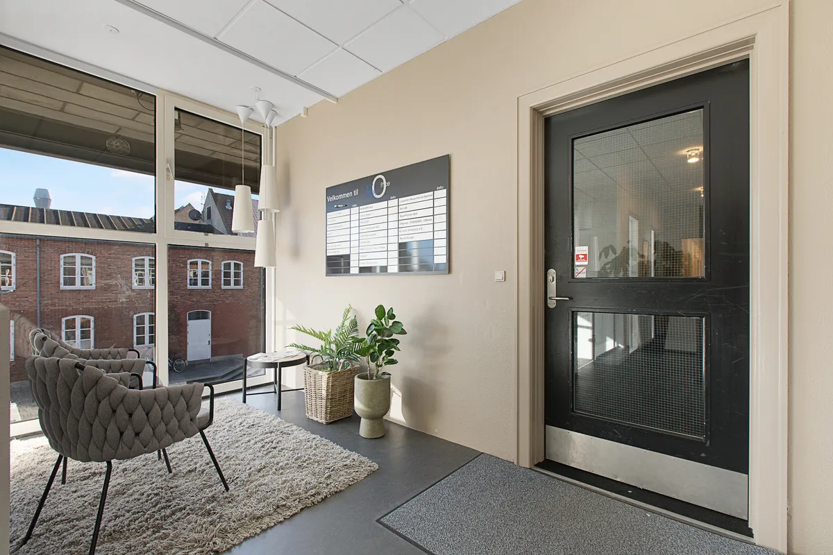 Kontor med indgangsdør og plakat. Et område med to stole, plante og tæppe. Vindue med udsigt til bygning. Lamper hængende