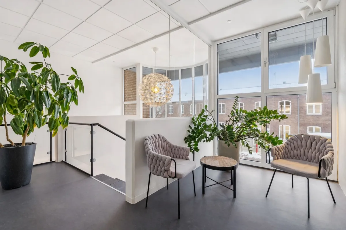 Kontorfællesskab med glasvinduer, lyse søjler og trægulve, plantepot, møbler og moderne lysekroner i hvidt og gråt