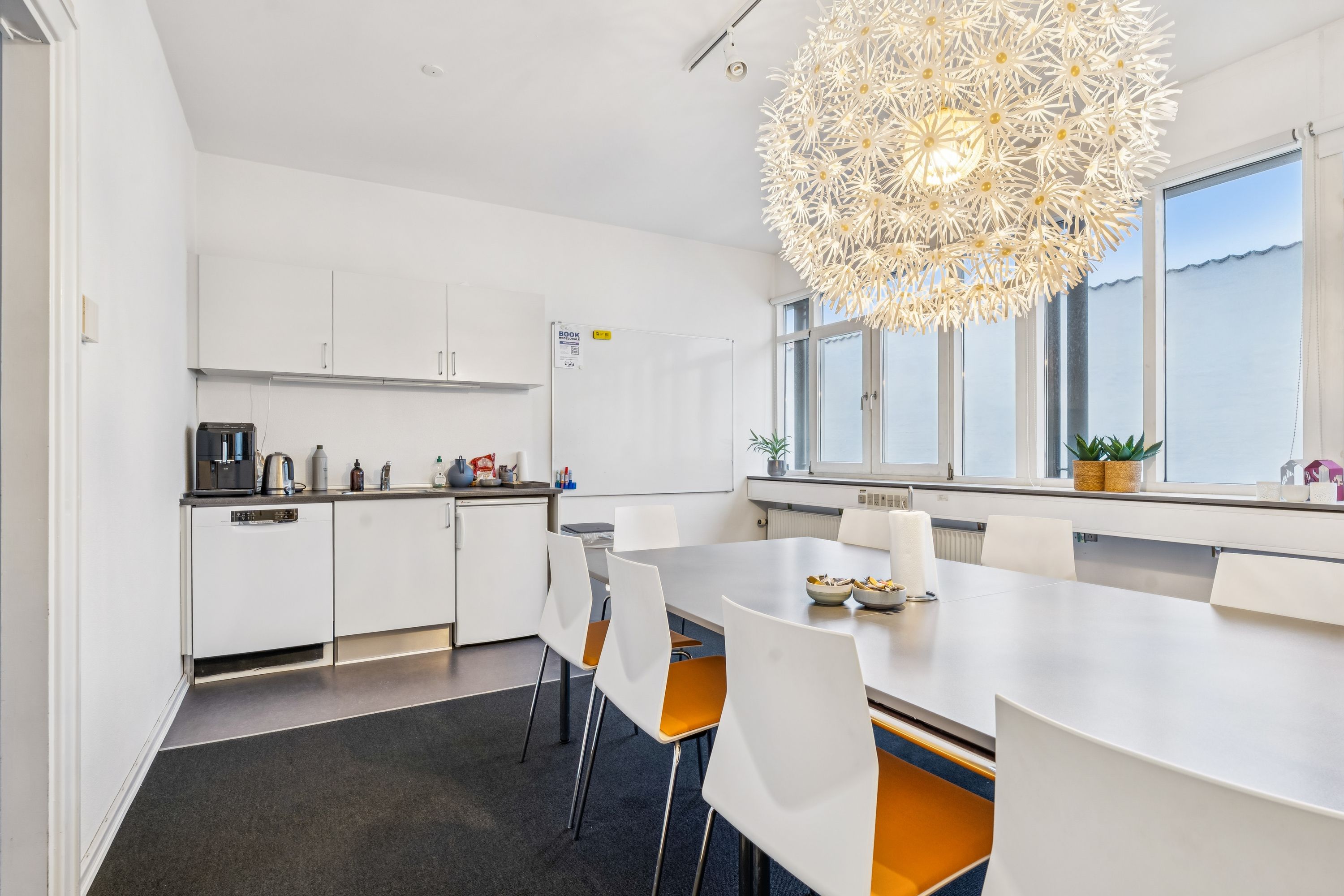 Kontorfællesskab med køkken, hvidt bord og stole, lysekroner, radiator, plantepot, kaffemaskine, køleskab, vindue og skrivebord med tavle og skrift