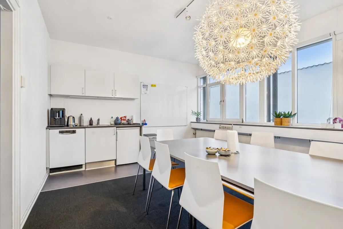 Kontorfællesskab med køkken, hvidt bord og stole, lysekroner, radiator, plantepot, kaffemaskine, køleskab, vindue og skrivebord med tavle og skrift