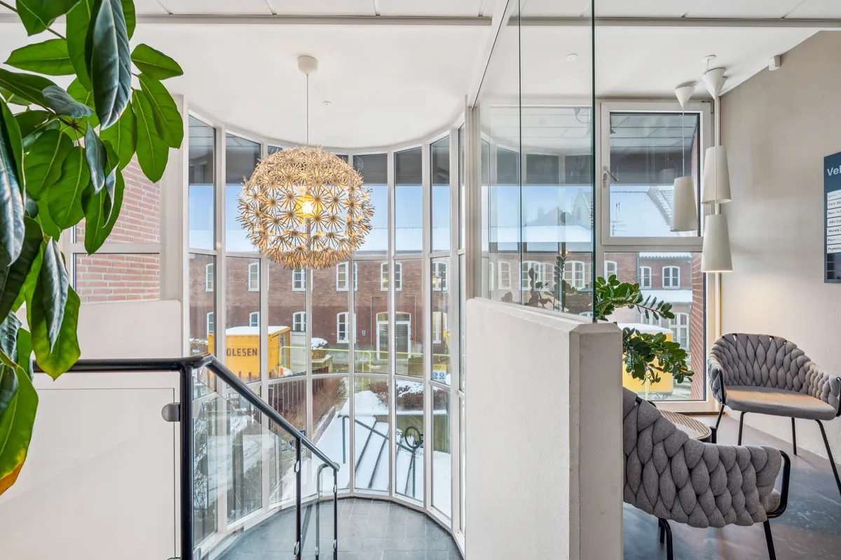 Kontor med lys og plante. Rundt gulv med glas og håndtag. Et stort gulvlampe og to sæder med ryglæneder. Udenfor er der en bygning med vindue og en vogn.