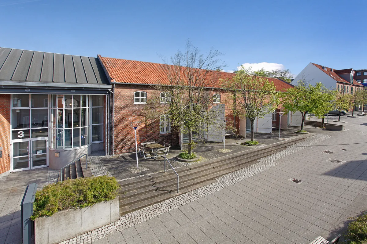 Kontorbyggeri med røde tegltag og glasvinduer samt terrasse med træer og møbler. Trapper med håndtag fører ned til gågade med brolægning.