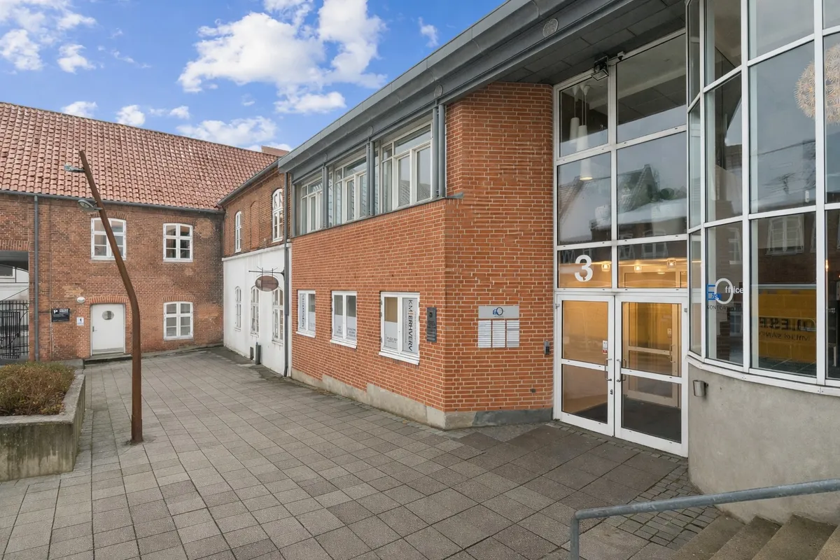 Et stort kontorblok med moderne design og store glasruder, nummer 3 ved indgangen, enkelte lysekroner i loftet og lyse, blå himmel med skyer