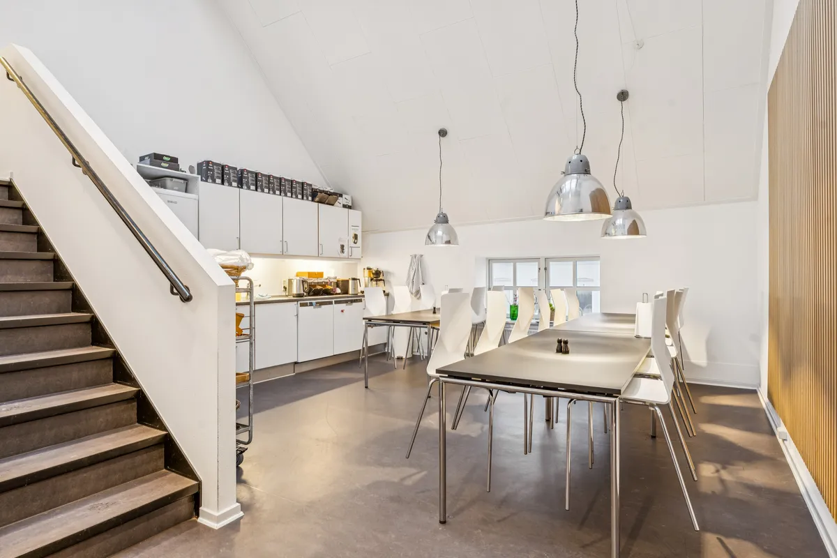 Kontorområde med to lange spiseborde og stole samt trapper til anden etage med håndtag og loftslampe