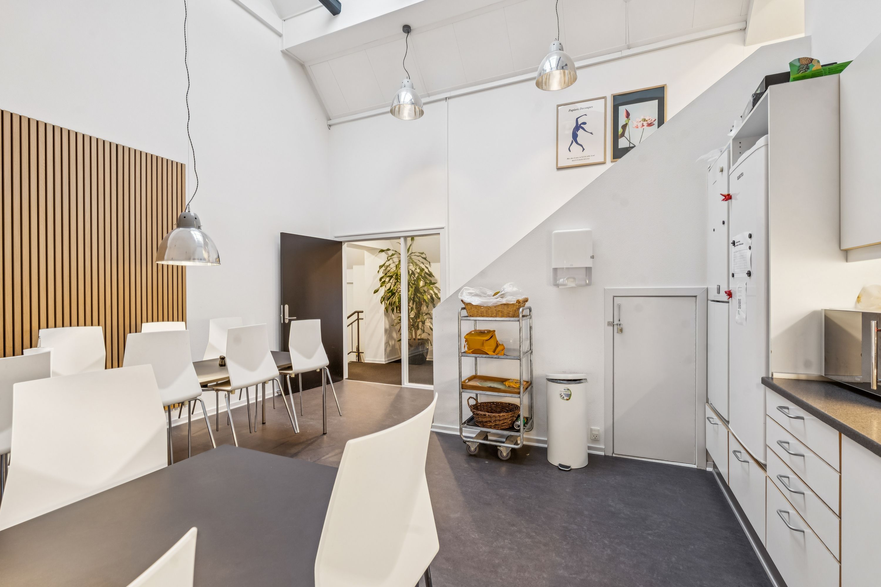 Kontorfællesskab med moderne design, hvide vægge, lysekroner, hvide stole, lyse bænke og en stor plante i baggrunden