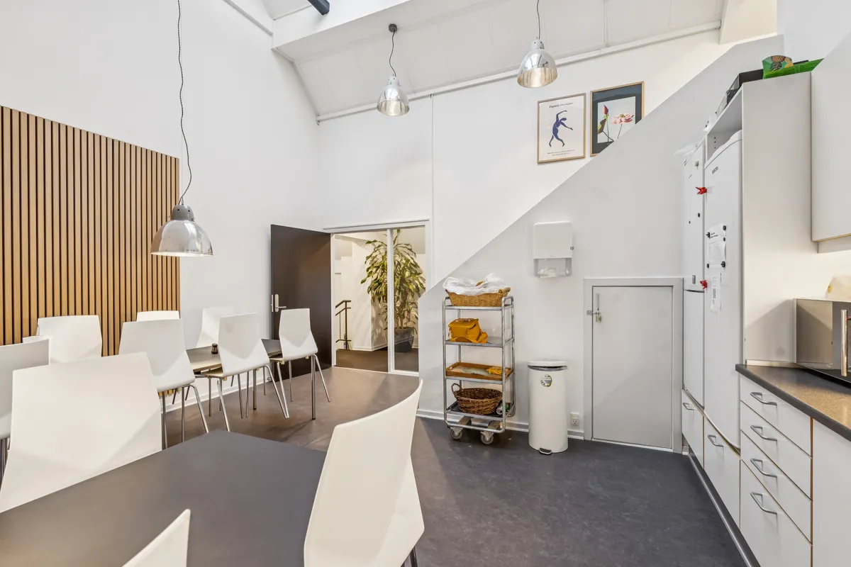 Kontorfællesskab med moderne design, hvide vægge, lysekroner, hvide stole, lyse bænke og en stor plante i baggrunden