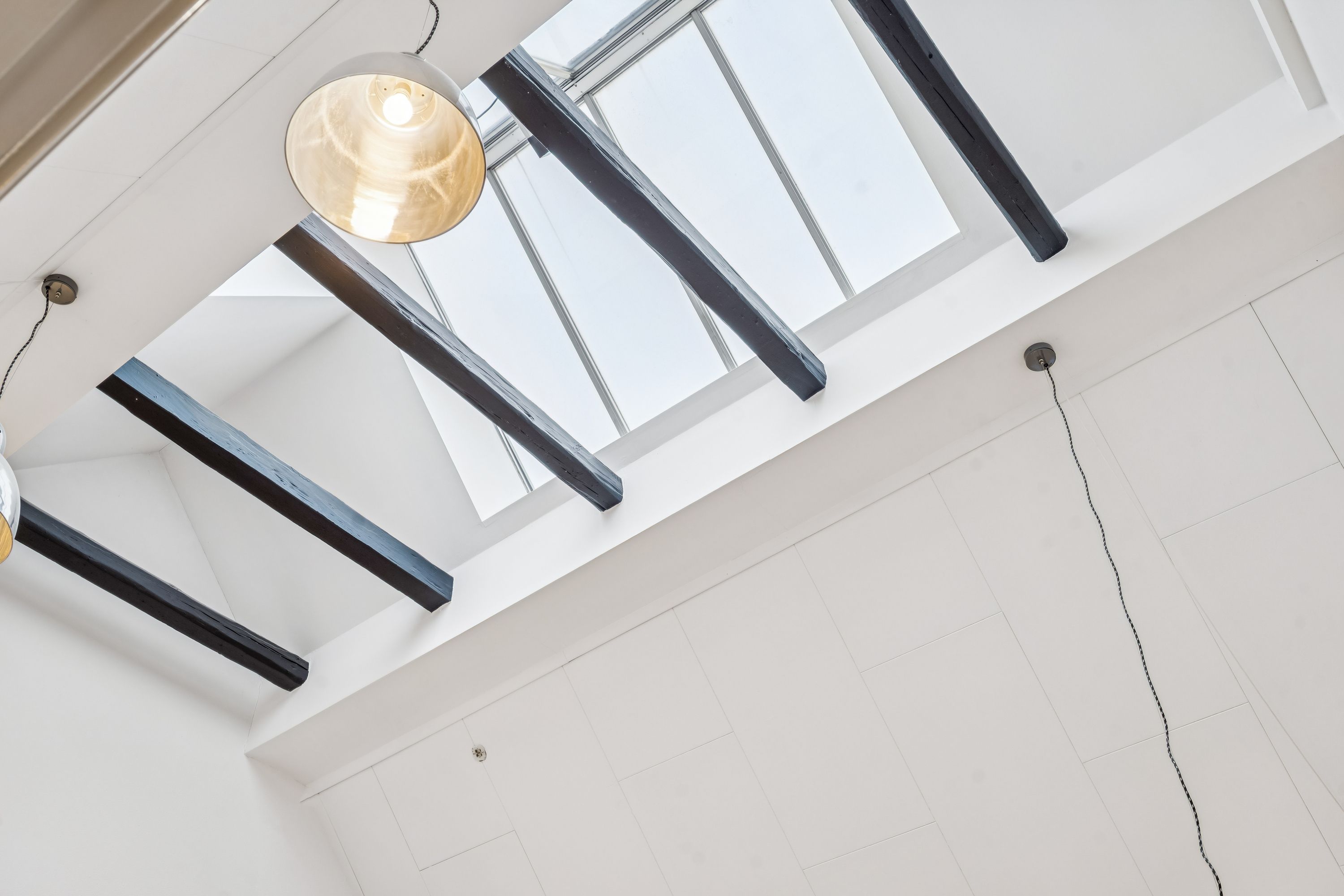 Loft med loftbjælker og loftsvindue, hvid forside og tre lysekroner hængende fra loftet, en med gult lys, to andre med sort ledning