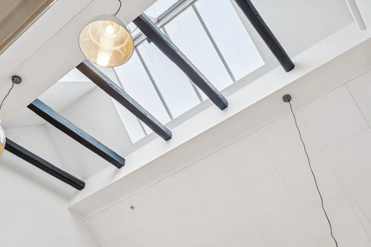 Loft med loftbjælker og loftsvindue, hvid forside og tre lysekroner hængende fra loftet, en med gult lys, to andre med sort ledning