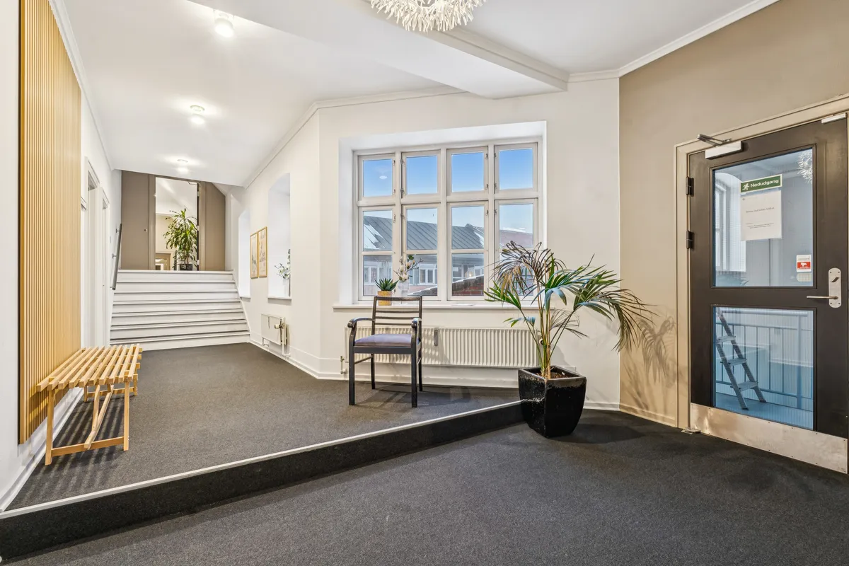 Kontorfællesskab med lyse vægge, vinduer og solrigt klima, møbler og plantager i indgangsområdet, radiatorer og trappe op til et loft