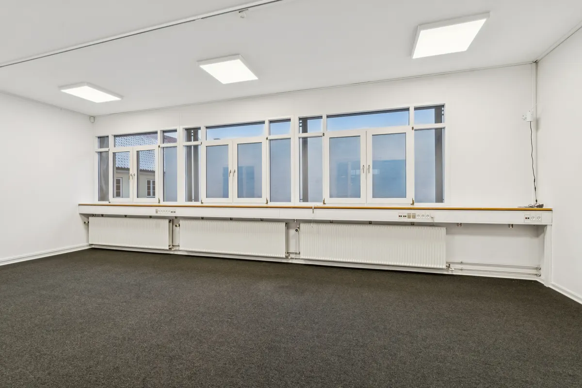 En tom og rummelig kontor med hvide vægge, lysekroner, radiatorer og vinduer med glasruder. En hvid elektrisk stikkontakt med stik er fastgjort til væggen