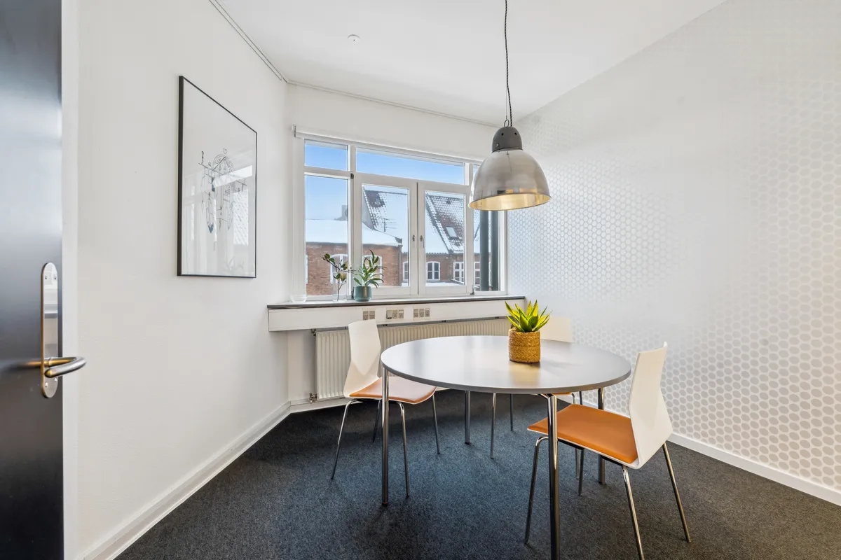 Et kontorfællesskab med et moderne møbelsæt og en rund bord med en plante i en plantekrukke på. Der er to køleskabe i vindueskarmen og en lampe hængende fra loftet