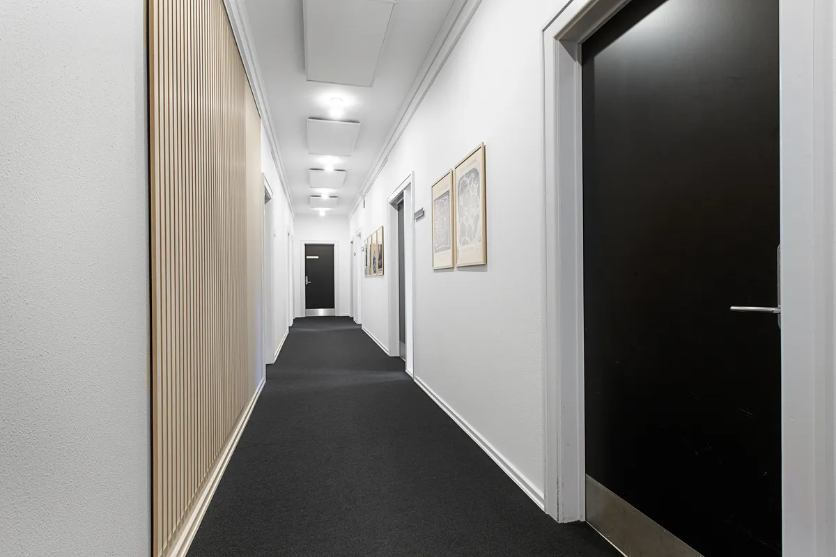 Kontorgang med hvide vægge og sortgule loftspaneler. Sortgule paneler og to malerier hængende på væggen. Sorte og hvide døre