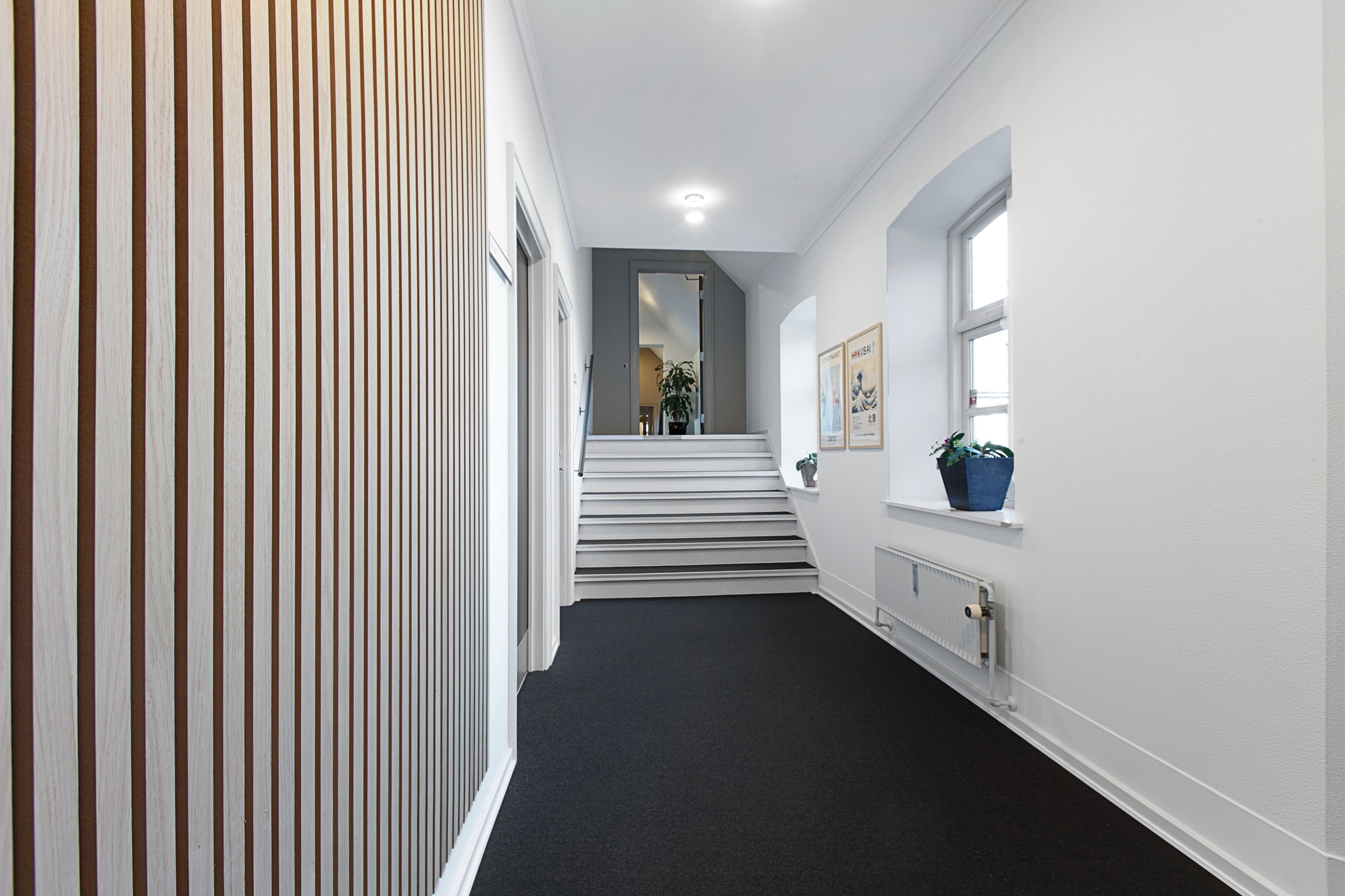 Inde i et kontorfællesskab med lyse vægge og møbleret gang. Et vindue med en plante og et maleri. Et hvidt trappeforløb og