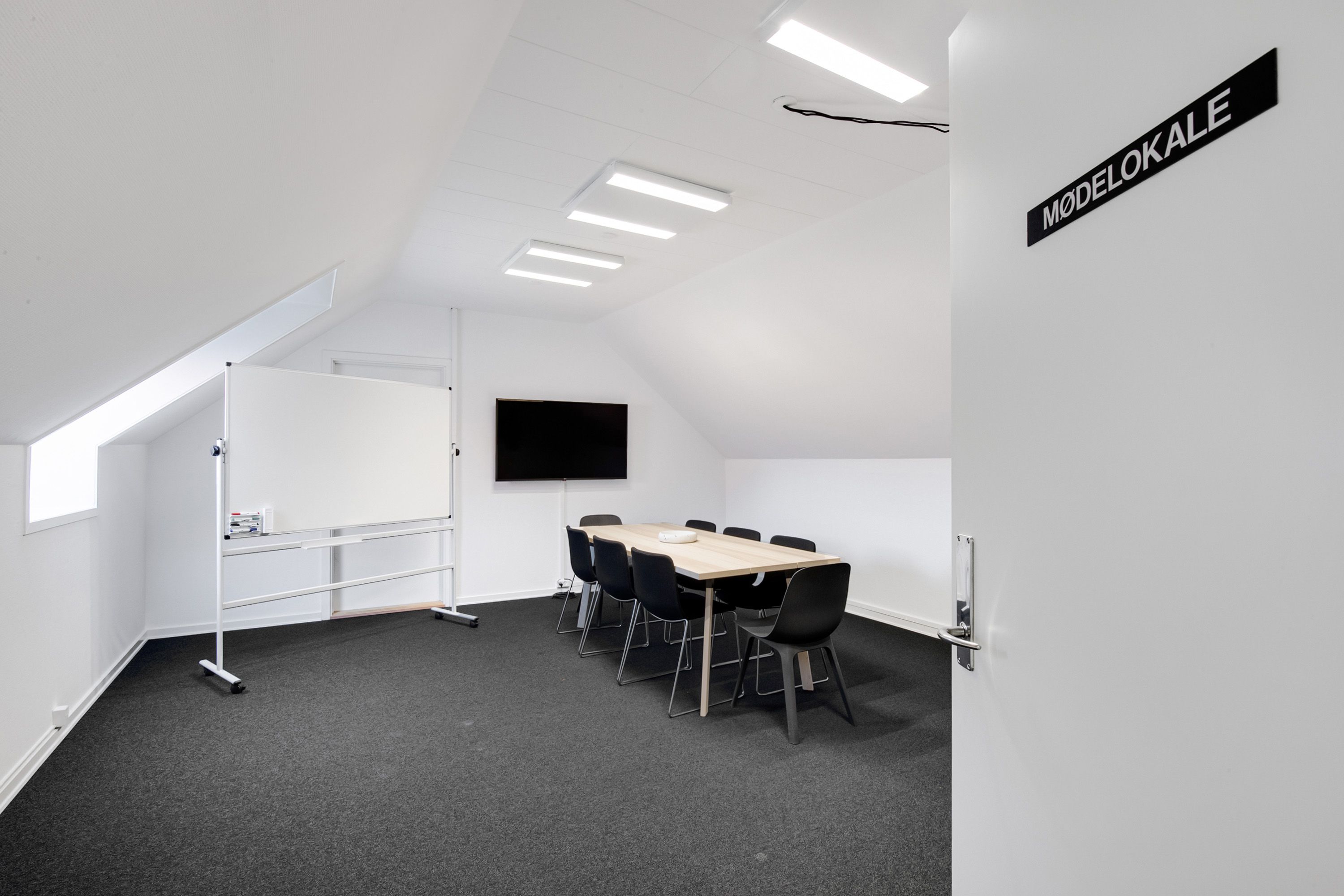 Et kontorlokale med hvide vægge og loft, lyseplader, et bord med stole, en whiteboard og en tv-skærm.