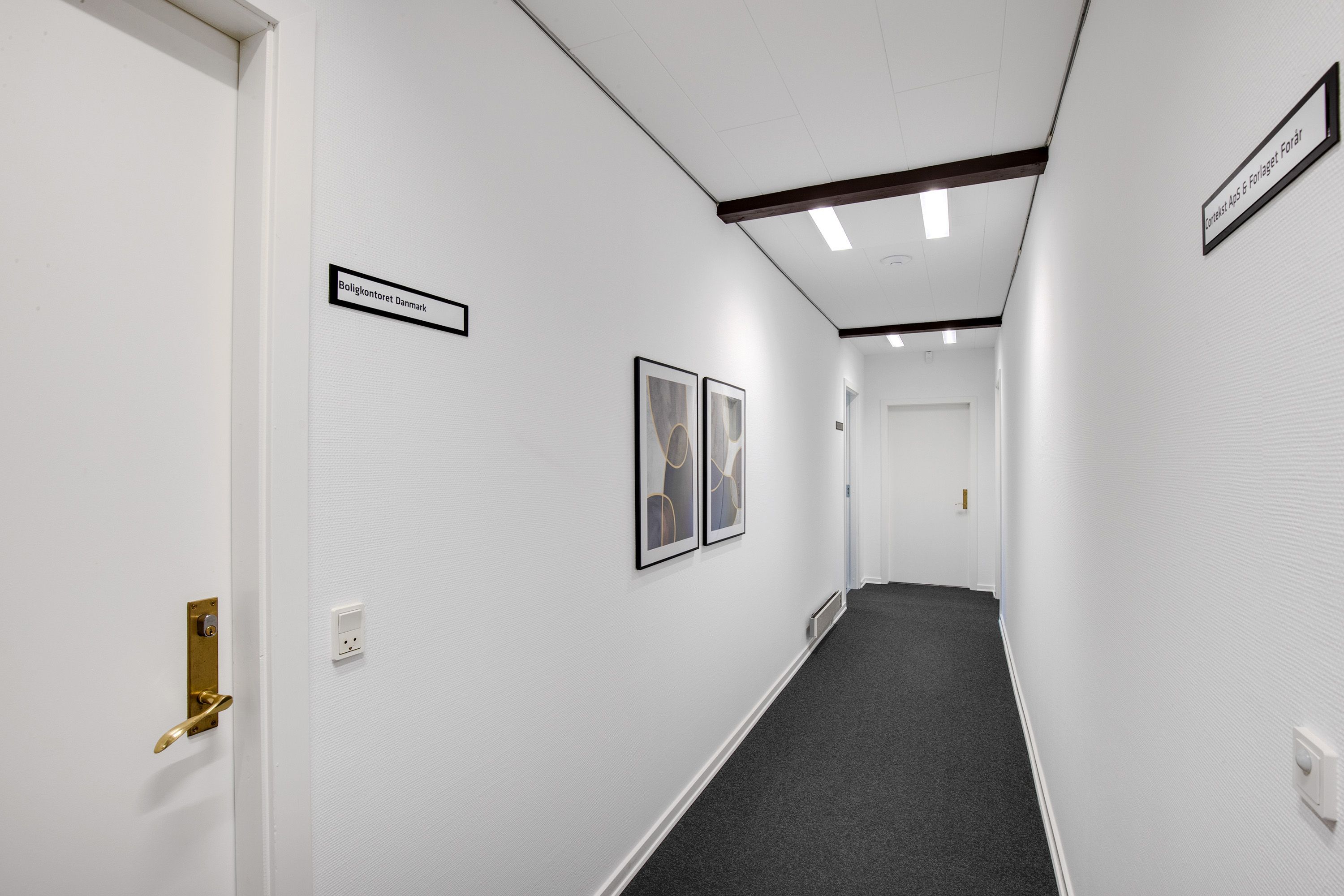 To moderne kunstværker hænger på en hvid væg. Der er to skilte med tekst. Gulvet er lige og hvide vægge er tydeligt synlige.