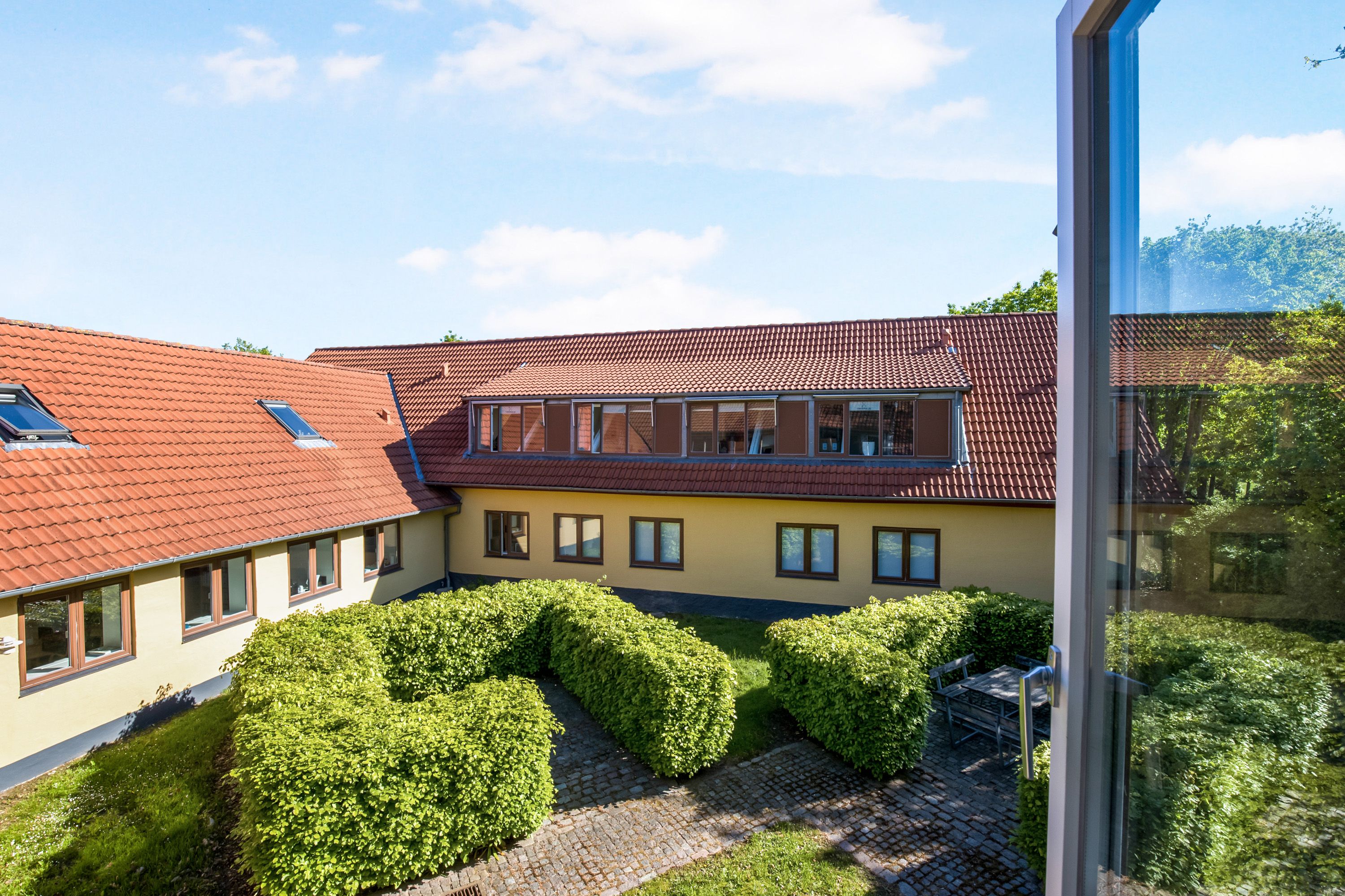 Bygning med rødt tag og lyseruder på facaden, der vender mod en have med skurvede buske og udsigt til et træ.