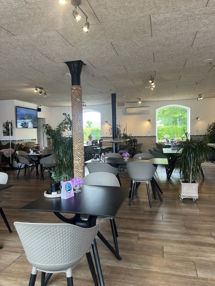 Restaurant med moderne møbler og høje lofter, to store vinduer med udsigt, lyse og varme farver, lysekroner, og et stort