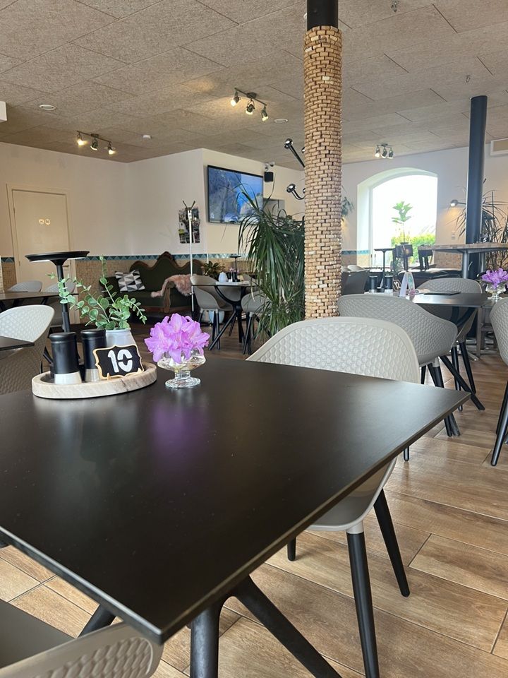 Et moderne indretning med lyse loftslamper og lysebrune gulvfliser, to borde med høje stole, og en plante med en vase med en