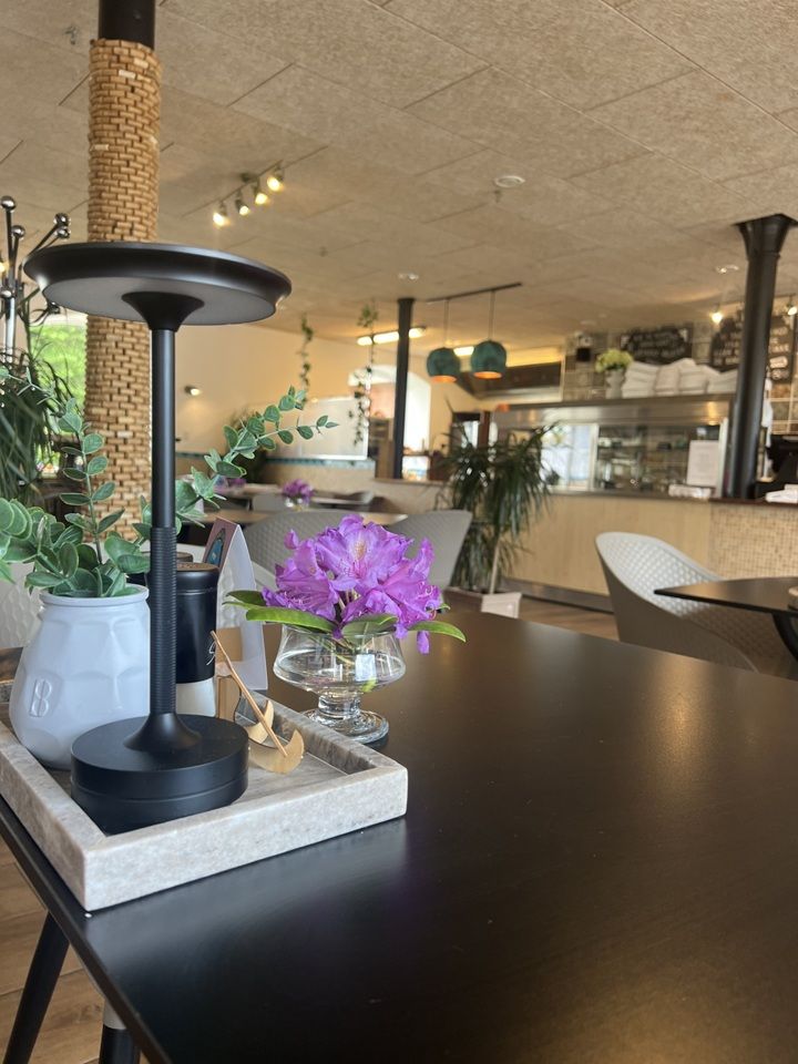 Sort bord med en vase med lilla blomster og en sort lampeskærm i et restaurantinteriør med trægulve og hvide stole