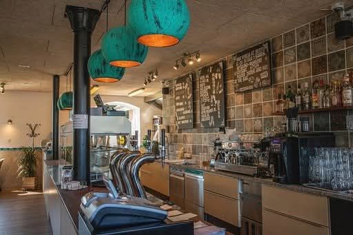 Billede af en restaurant med trægulve, lyse lysekroner og køkkenudstyr. Der er borde og stole og en kaffemaskine.