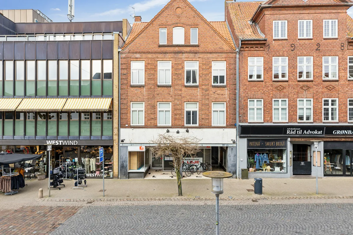 På en gade er der en bygning med flere butikker og kontorlokaler, der tilbydes til leje. Der er træ og cykler parkeret