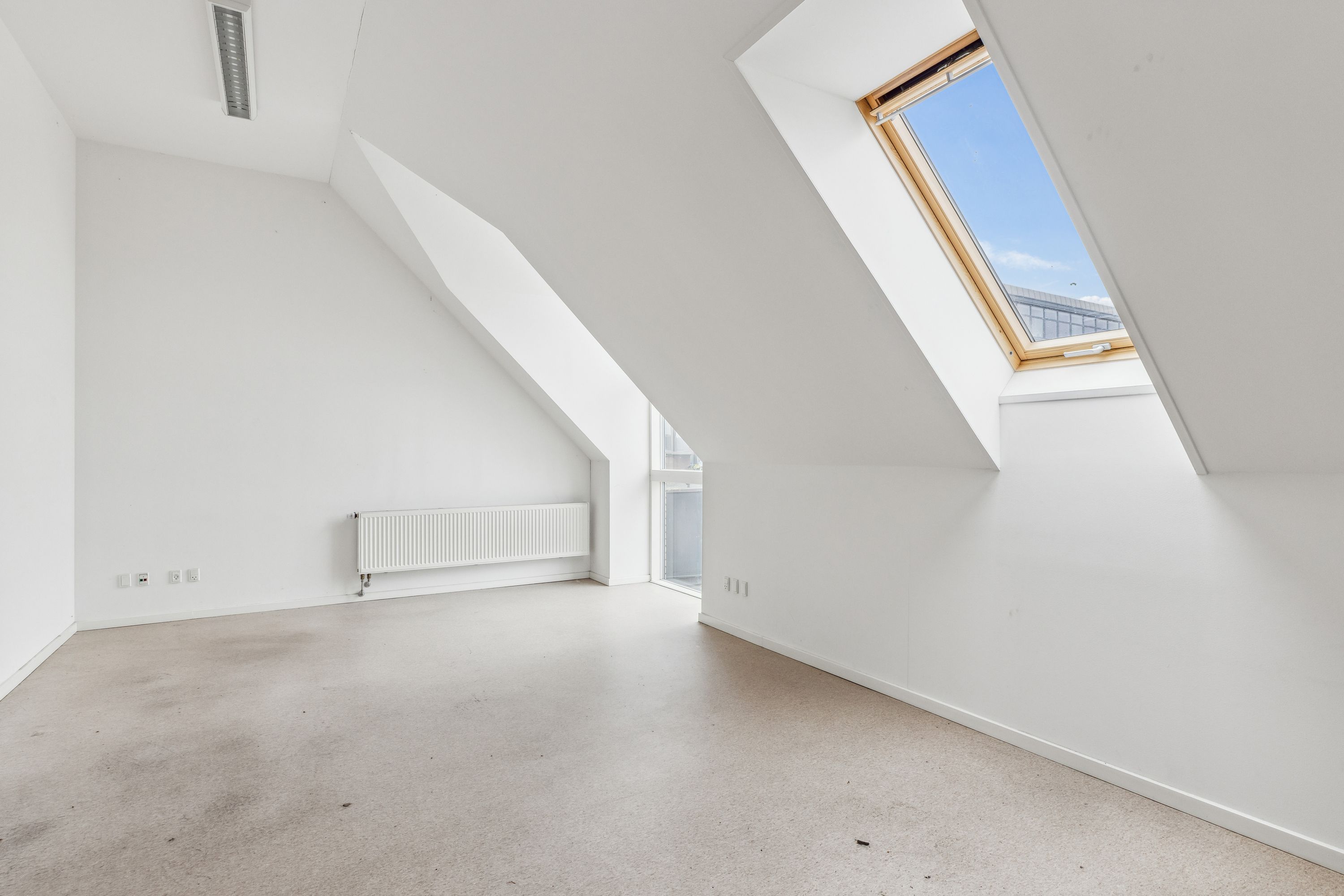 En tom, hvid, lyse loftsværelse med lysegul gulvbelægning og træruderede loftsvindue med udsigt til en bygning i baggrunden