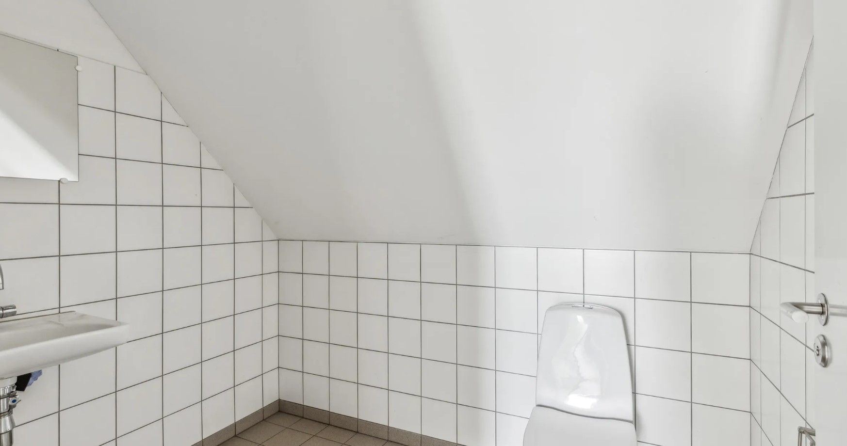 Kort kontorlokale med lysegrå flise på gulvet og hvide flise på væggene med toilet, spejl og vask