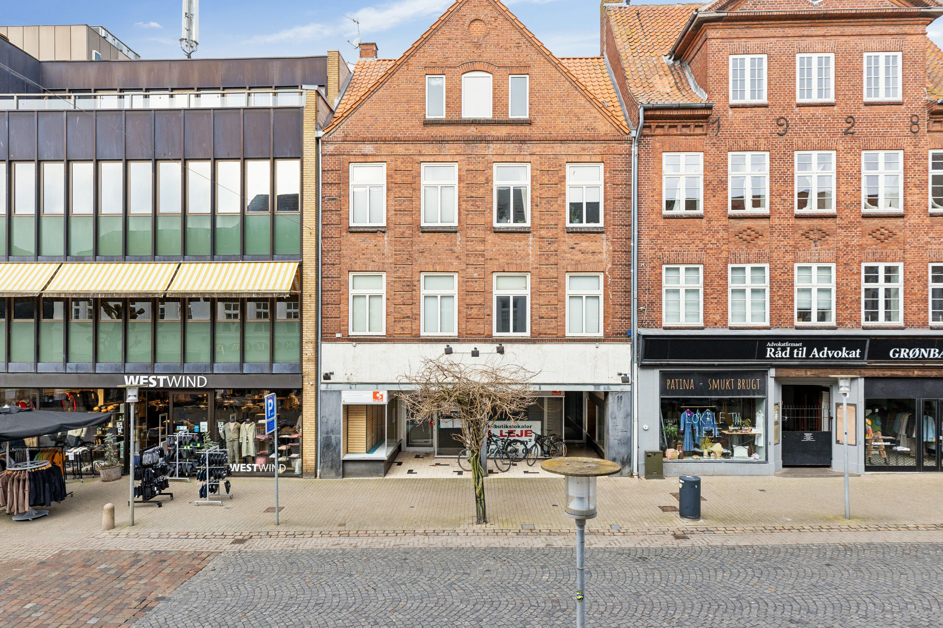 En bred gade med gamle og moderne bygninger, hvor en række butikker er placeret. Butikkerne inkluderer Westwind, Patina