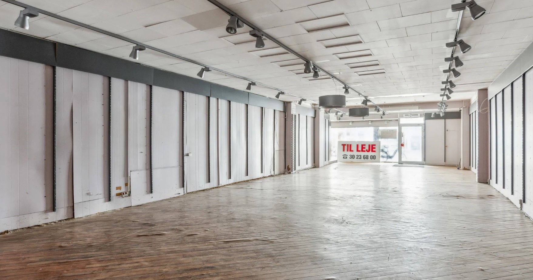 Kortere række af tomme butikker med hvide vægge og lyse lofter med sporlys og et skilt med teksten Til Leje.
