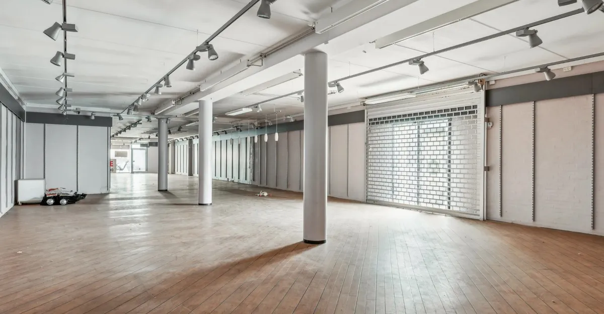 Store tomte, gulve med træbelægning, højloft og gulvmonterede søjler, hængende lamper, hvide vægge og en åben garagedør med