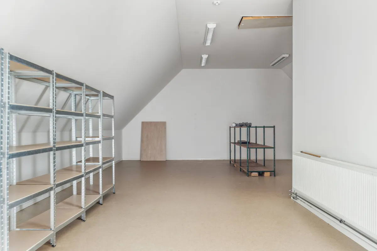 En tom stue med metalhylder, to brædder, tre hvide loftslamper og en hvid radiator på højre side. Gulvet er lavet af let at