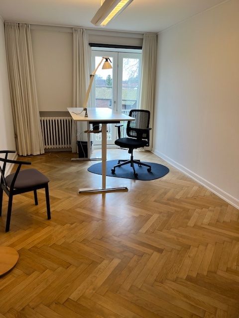 Kontor med højloft, højloftsvinduer, sort stol, bord, gulvbelægning og hvide vægge med gardiner. Lys og stol står på gulvet.