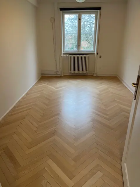 Hvidt værelse med lyse trægulve og gulvbelægning i stribet mønster. Væggene er hvide og rummet har et vindue med en radiator