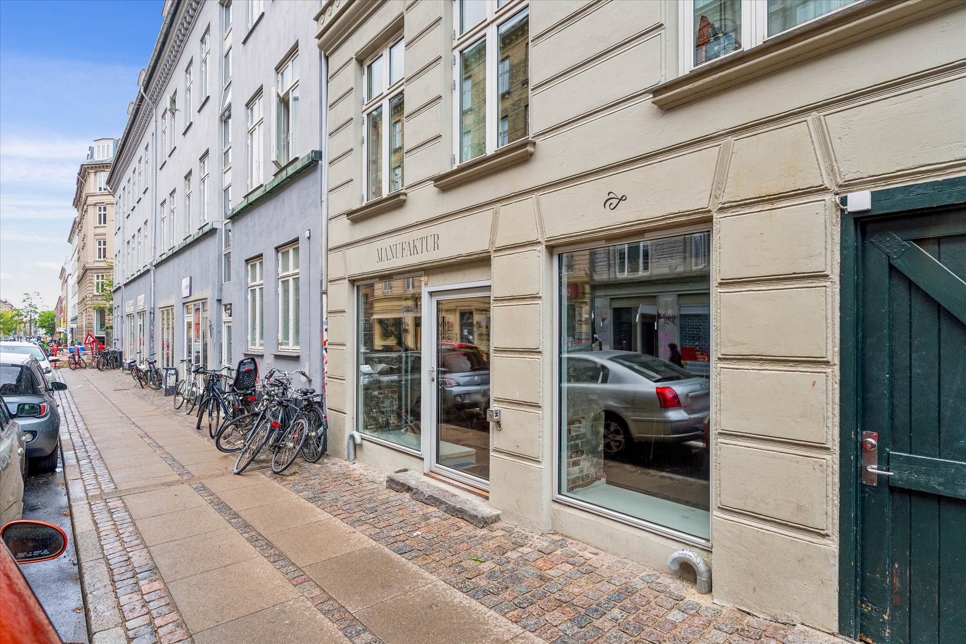 En facade med butikker og kørende biler på Griffenfeldsgade, Nørrebro. En bil reflekteres i butikkens ruder og der er mange