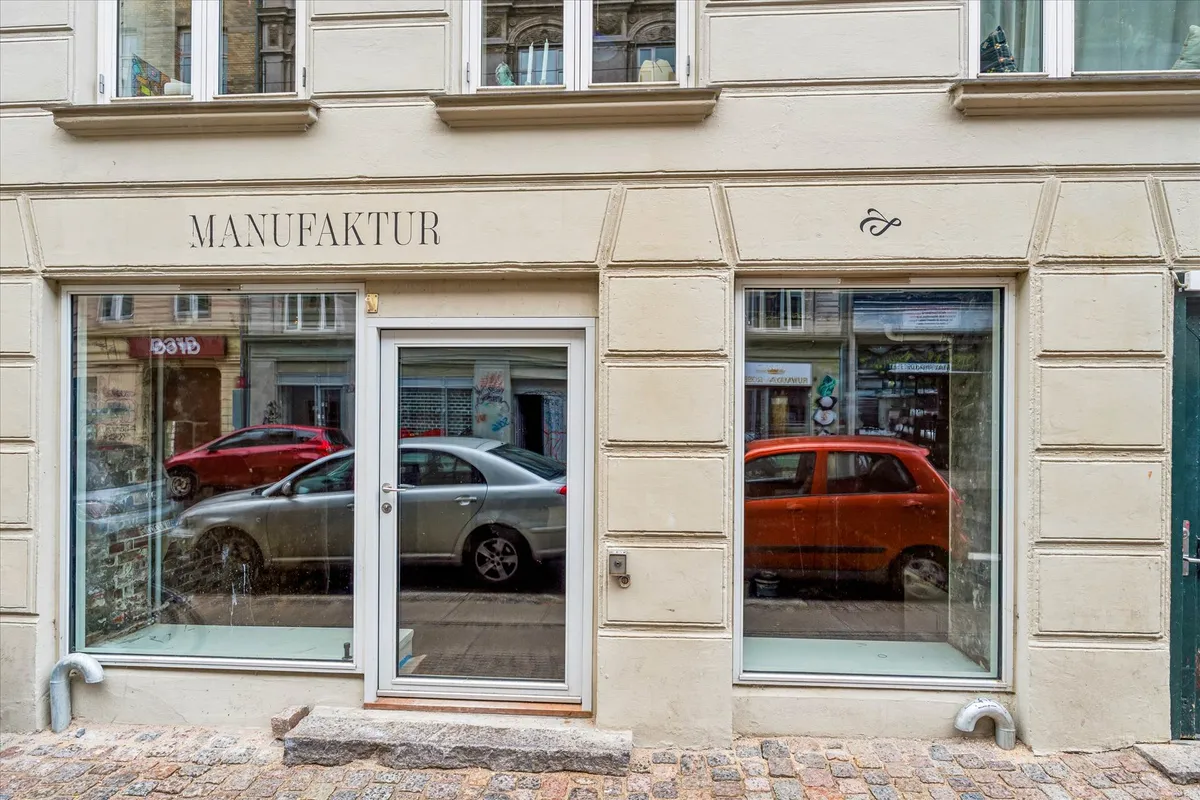 Et lille butiksvindue med teksten Manufaktur i toppen og en refleksion af to biler på hver side af vinduet