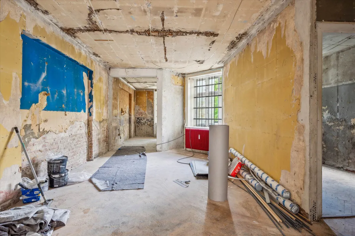 Råt erhvervslejemål med revner og skrællet maleri. Tømmer og rør på gulvet og søjle med rødt radiator.