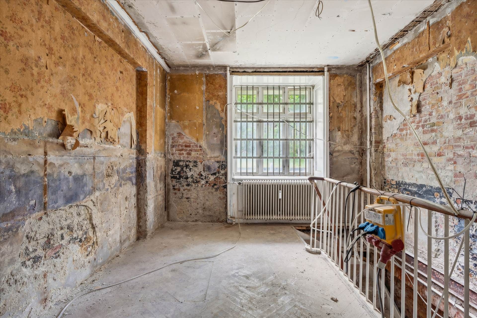 Råt og forfaldent erhvervslejemål med tomme vægge, gammel radiator, vinduesgitter og skadet gulv og loft