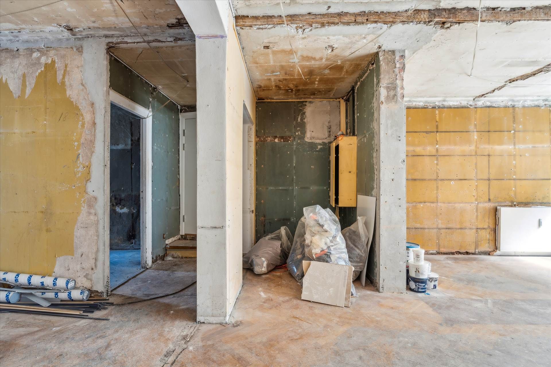 En renoveringszone med trægulve, søjler og skade på vægge og loft. Stykker og poser er samlet på gulvet.