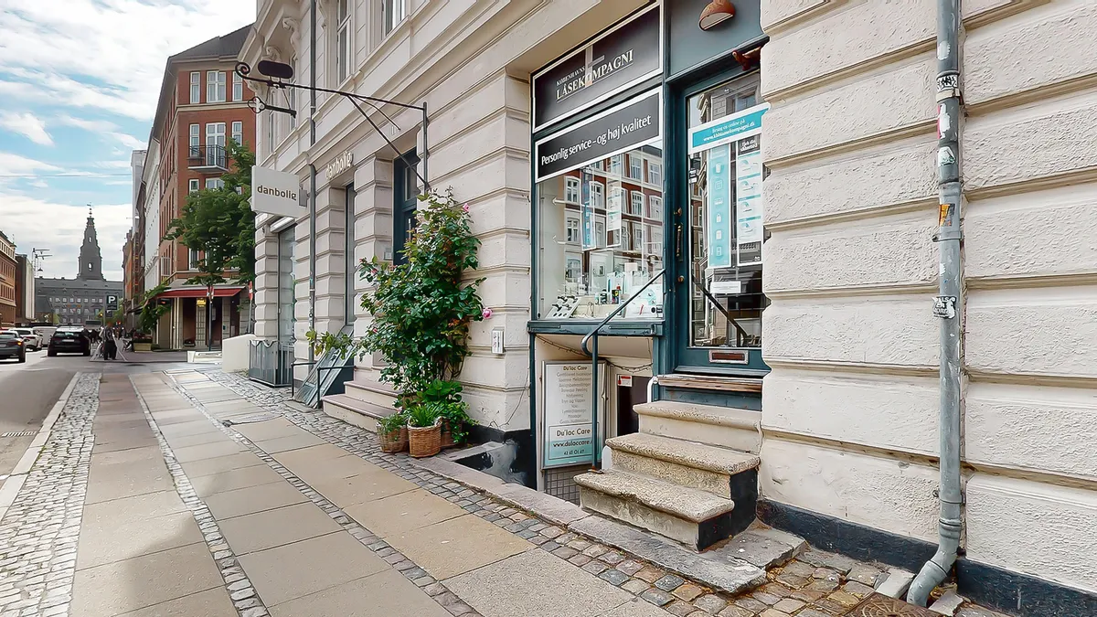 En butik med røde ruder og grønne planter udenfor. Der er skilte med tekst og logoer. Stigene fører op til indgangen, og der