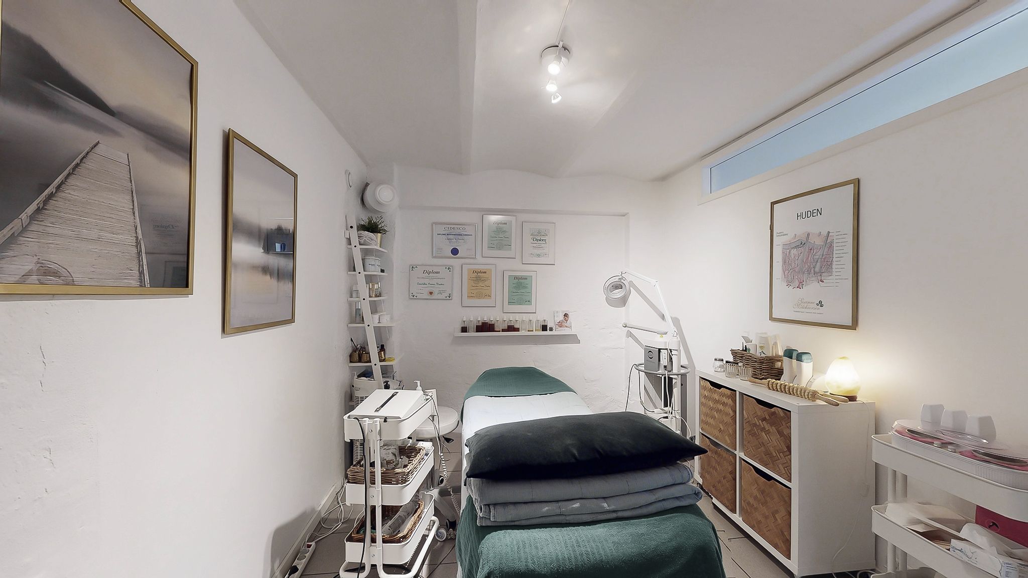 Behandlingsrum med massagebord, hylde med certifikater og plante samt lysarmatur på loftet og lyse puder og tæpper på sengen