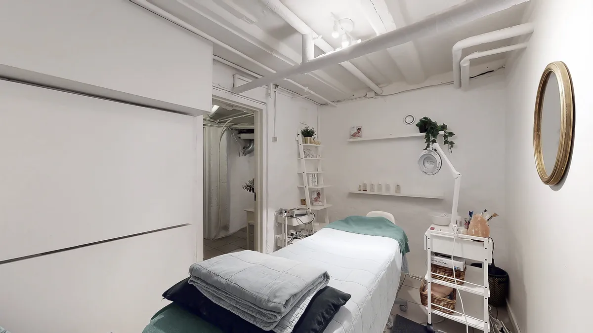 Hjørne med massagebord og grønt tæppe samt flere hvide højreokker med plante. Hvid væg og plante i guldfarvet spejl