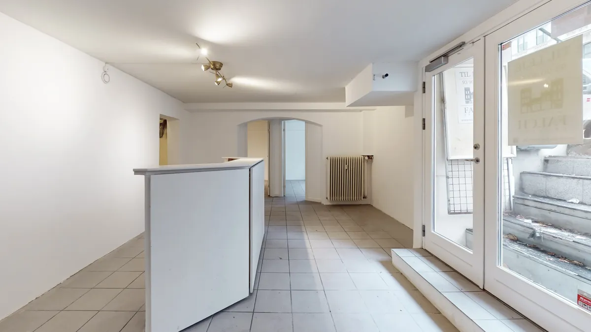 Indgangsområde til lejelejlighed i København K med moderne hvide vægge og gulv samt lysekroner og skrivebord. Stiger udenfor og radiator mod bagvæggen.