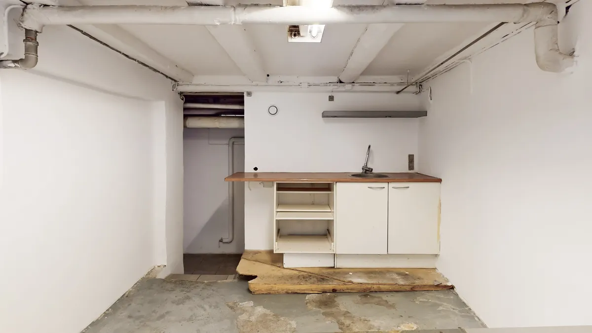 Et tomt kælderrum med et køkken og en dør i baggrunden, hvor gulvet og væggene er fugtet og svært beskadiget.