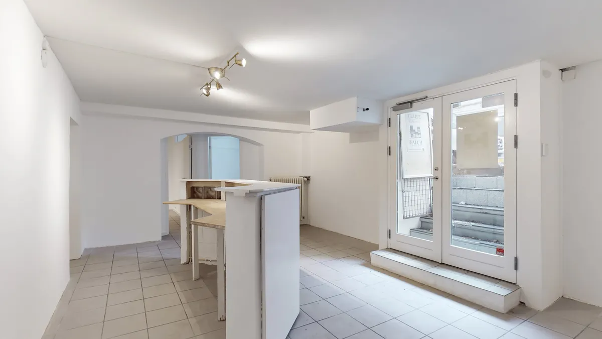 Moderne og lyse lokaler med hvide vægge og gulv, hvilket giver en minimalistisk følelse. Der er et slankt, hvidt kontorbord og en høj bar.