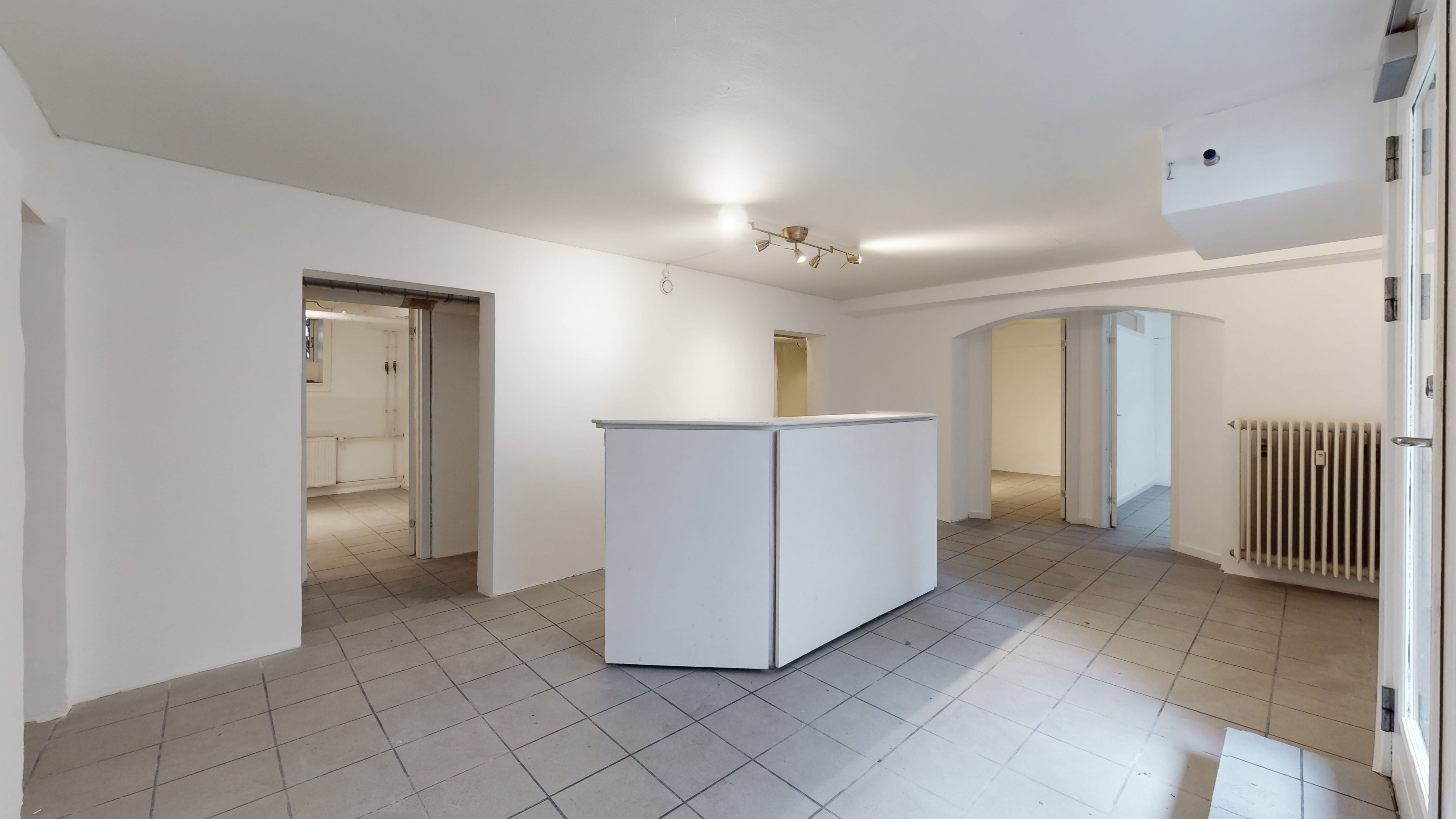 En stor, ren, hvid stue med gulvbelægning og tre døre. Væggene er hvide og der er en radiator i højre side af rummet. Der er en lyskrone og et kamera på loftet.